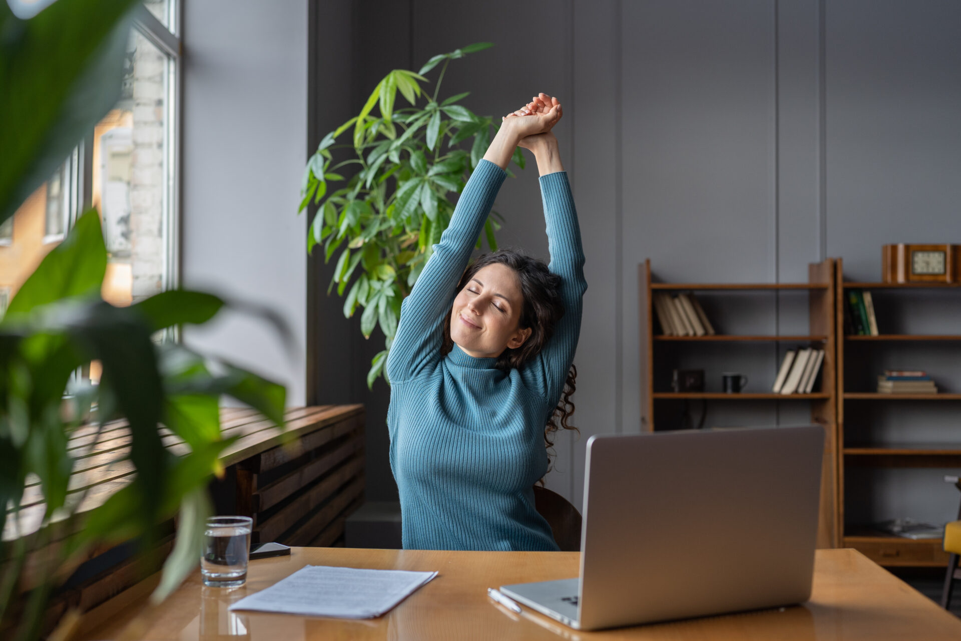 A woman sits at a desk with a laptop, glass of water, and papers and stretches her arms up into the air.