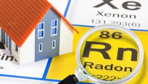 The radon symbol is shown on a periodic table under a magnifying glass next to a small toy house figure.