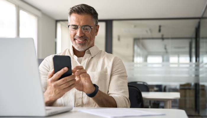 A smiling man sits at a table scrolling on a black smartphone. A laptop sits open on the table next to a pile of papers.