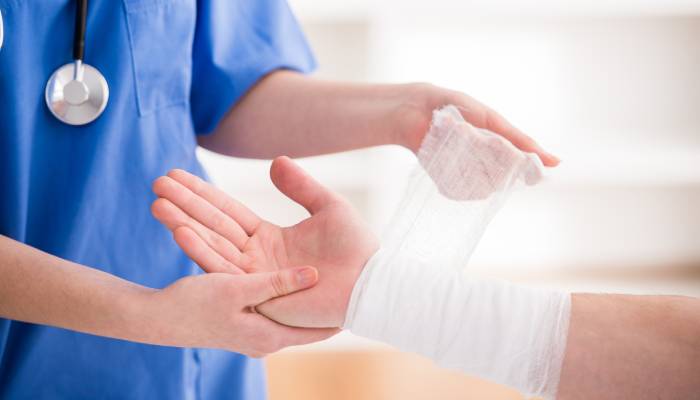 An individual wearing blue scrubs and a black stethoscope is applying a white wound dressing to a person's arm.