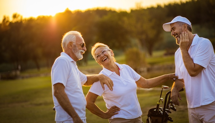 Three senior adults wearing white shirts stand on a golf green while talking and laughing as the sun sets.