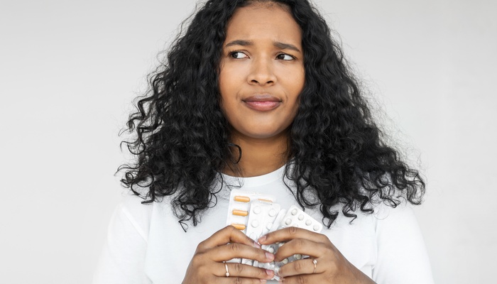 An African American woman with curly hair holds various pill packs with an uncertain facial expression.