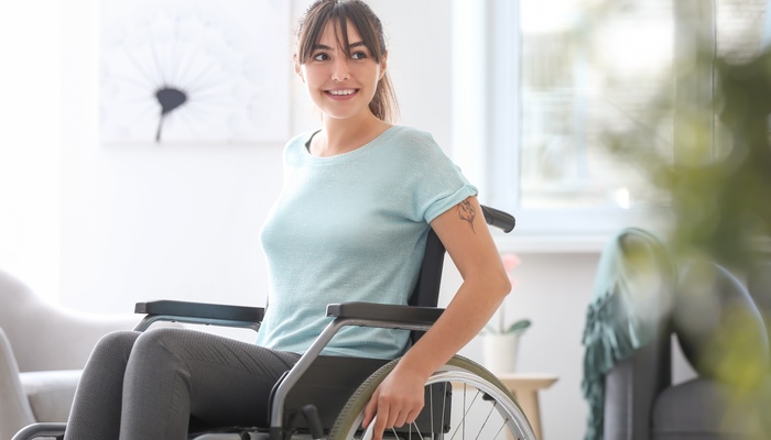 A young woman with brown hair sits in her wheelchair in her home. She looks to the side and smiles.