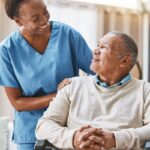 A Black woman nurse is smiling and taking care of an older man, who is wheelchair bound. They're in a living room.