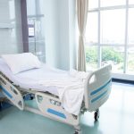 An empty hospital bed contains a white pillow and a white blanket. A blue nightstand sits next to the bed.