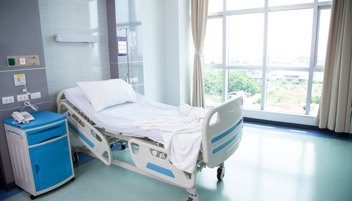 An empty hospital bed contains a white pillow and a white blanket. A blue nightstand sits next to the bed.