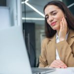 A woman is working on her laptop at work, with a toothache. She's clutching the side of her face in pain.
