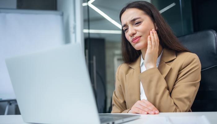 A woman is working on her laptop at work, with a toothache. She's clutching the side of her face in pain.