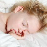 A close-up shows a small child sleeping on a white sheet, sucking their thumb. A white blanket covers the child.