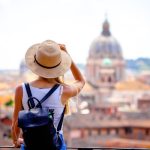 A woman stands on a balcony overlooking the city of Rome, Italy, during the daytime. She wears a backpack.
