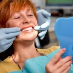 A red-headed senior woman leans back in the dentist chair as she holds a mirror up to her denture smile.