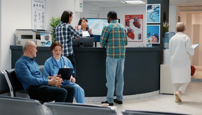 A busy hospital emergency room reception desk area. Patients sit and stand in the waiting area talking to nurses.