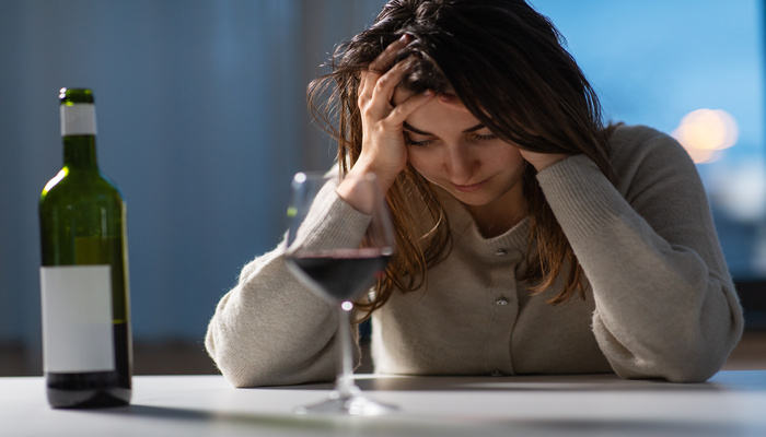 A young woman sitting at a bar with her head in her hands. A glass of red wine and the bottle rest in front of her.
