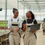 Two women walking together through a greenhouse farm. One of them is holding a laptop while they're talking.