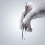 A close-up of a hand wearing white latex gloves. The hand holds stainless tweezers down against a gray background.