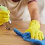 Home 2 Someone wearing yellow gloves sprays a cleaning chemical onto a wooden counter. They use a blue towel to wipe it up.