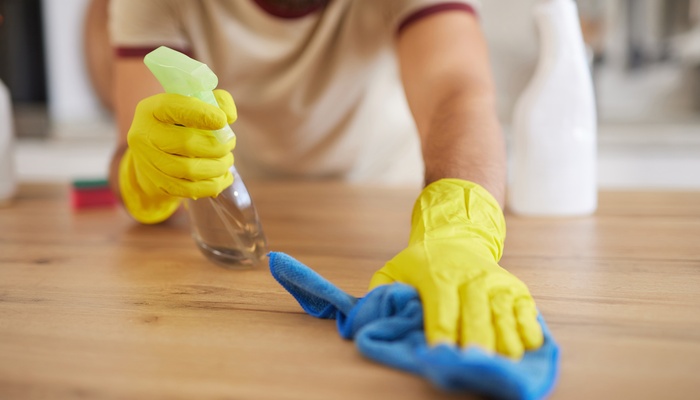 Someone wearing yellow gloves sprays a cleaning chemical onto a wooden counter. They use a blue towel to wipe it up.