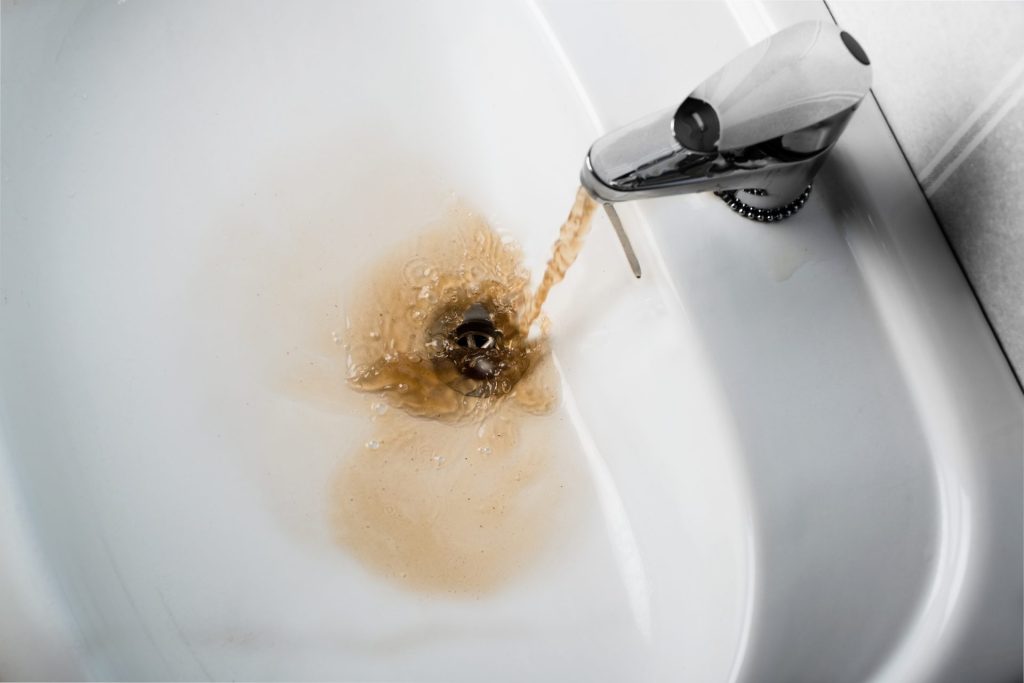 Reddish-orange, rusty water flowing from a stainless steel faucet into a clean white sink and going down the open drain.