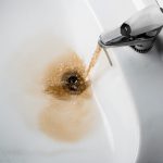 Reddish-orange, rusty water flowing from a stainless steel faucet into a clean white sink and going down the open drain.