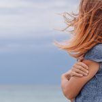 Home 5 A young woman stands alone outside on a cloudy day. She's in a gray T-shirt, and the wind blows her hair over her face.