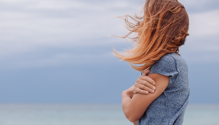A young woman stands alone outside on a cloudy day. She's in a gray T-shirt, and the wind blows her hair over her face.
