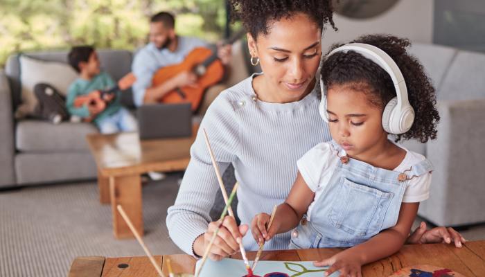 A mother and daughter work on a painting in the foreground. A father and son play guitars in the background.