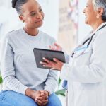 Home 2 A woman sitting on a hospital examination bed while speaking with a doctor. The doctor is holding a tablet.