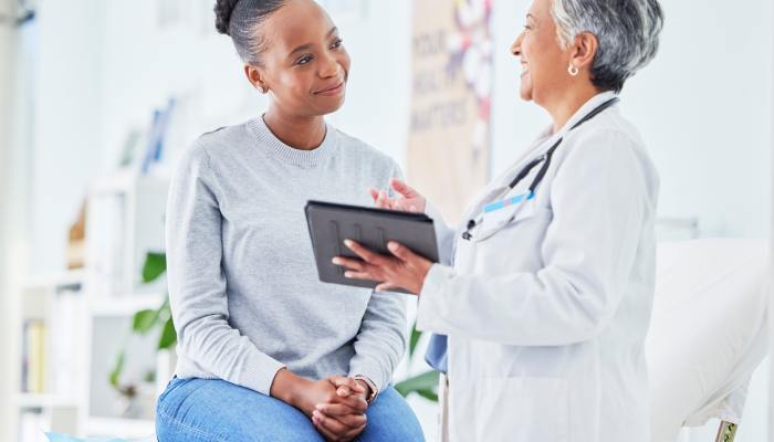 A woman sitting on a hospital examination bed while speaking with a doctor. The doctor is holding a tablet.