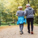 An older man and woman in athletic wear walk side-by-side on an outdoor path surrounded by tall trees.