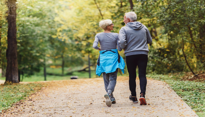 An older man and woman in athletic wear walk side-by-side on an outdoor path surrounded by tall trees.