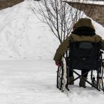 A person in a wheelchair facing a large snow pile against a brick wall. The person wears a green winter jacket and gloves.
