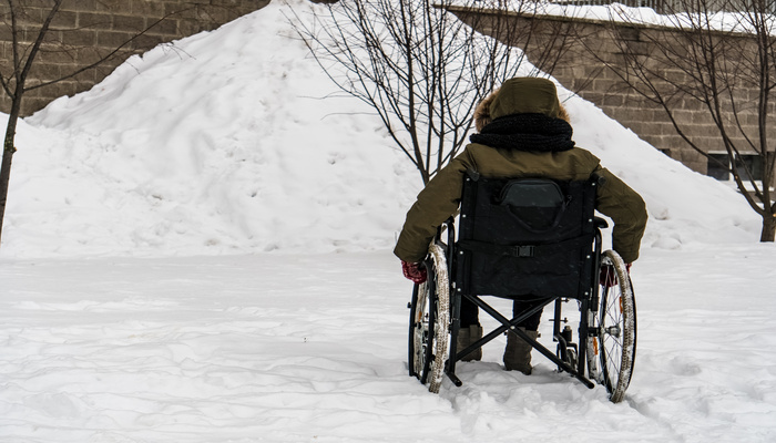 A person in a wheelchair facing a large snow pile against a brick wall. The person wears a green winter jacket and gloves.