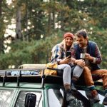 Home 3 Two people sitting on a green truck in the woods. One person holds their phone, and another person holds a metal mug.