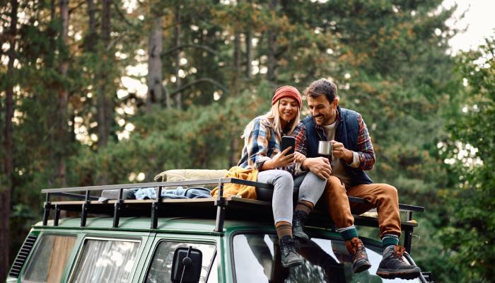 Two people sitting on a green truck in the woods. One person holds their phone, and another person holds a metal mug.