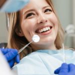 A woman wearing a dental bib smiles widely. A blurred dentist sits in front of her holding dental instruments.