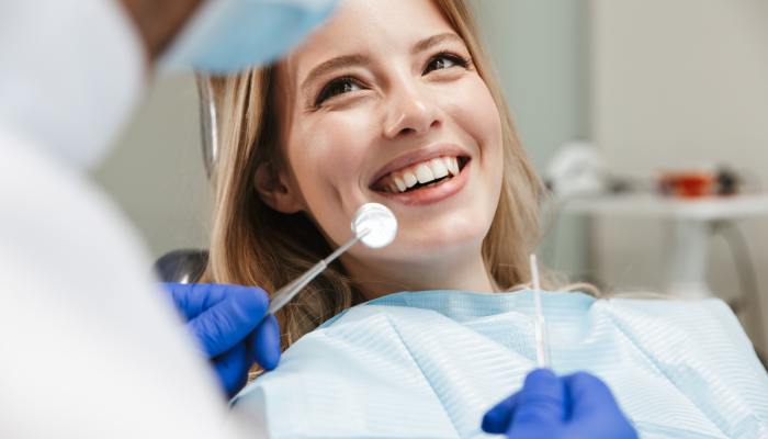 A woman wearing a dental bib smiles widely. A blurred dentist sits in front of her holding dental instruments.