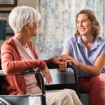 An older woman sits in a wheelchair in her living room. A young-adult woman sits next to her on a couch and holds her hand.