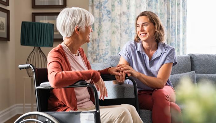An older woman sits in a wheelchair in her living room. A young-adult woman sits next to her on a couch and holds her hand.