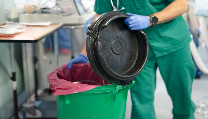 A doctor wearing a smart watch and green scrubs is opening a biohazard medical waste container in a hospital room.
