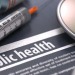 A close-up of a piece of paper that says 'public health' in all caps next to a stethoscope and needle.