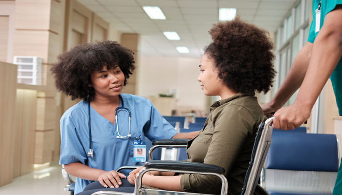 A person wearing scrubs kneels down to talk to another person who is sitting in a wheelchair in a healthcare facility.