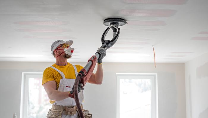A man in a t-shirt, overalls, hard hat, and mask sanding drywall on the ceiling of a home with a sander.
