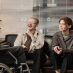 Home 3 A man and a woman smiles while sitting on a black couch, listening to a man with a disability in an office setting.