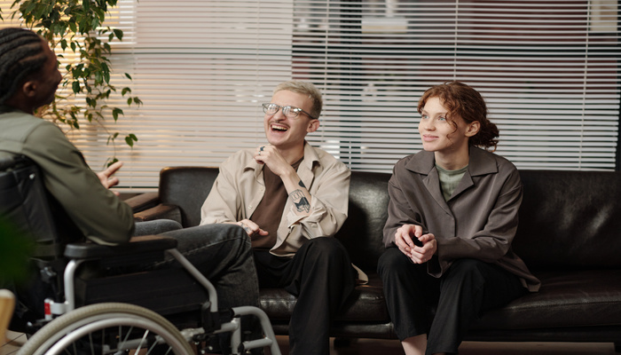 A man and a woman smiles while sitting on a black couch, listening to a man with a disability in an office setting.