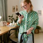 Home 2 A young woman standing next to her desk through the use of a walker mobility aid. She is looking at her smartphone.