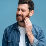 Home 2 A man grimacing, with teeth gritted, and holding a hand up to his cheek on the right side of his face.