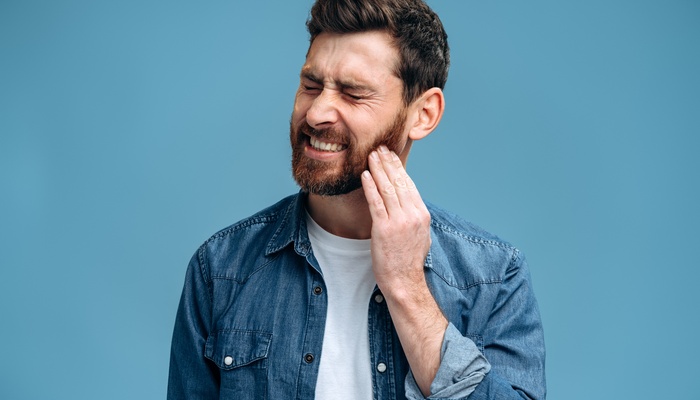 A man grimacing, with teeth gritted, and holding a hand up to his cheek on the right side of his face.