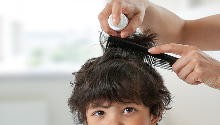 A close-up of a child's head with a person off-screen combing through his black hair and holding a product bottle up to spray it.