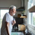 Home 5 A senior man stands at his kitchen sink with a glass of water as he stares out of his kitchen window.