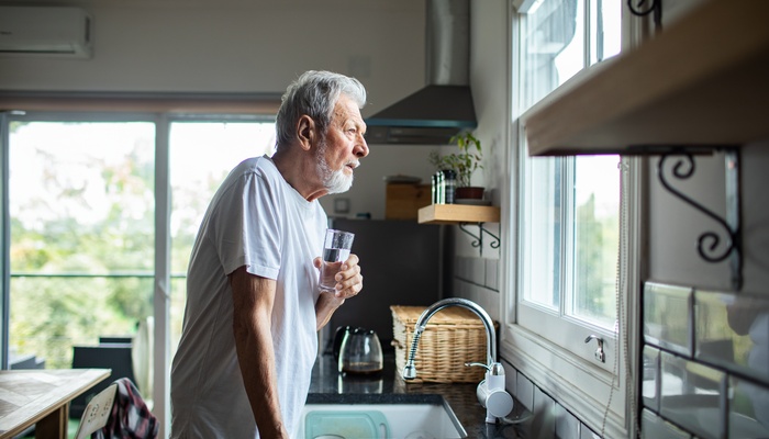 A senior man stands at his kitchen sink with a glass of water as he stares out of his kitchen window.